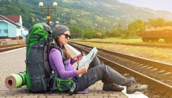 Happy girl tourist with backpack sits on a platform waiting for a train. Travel concept. Traveler holding map, waiting for a train and planing for next trip. Traveler reading map